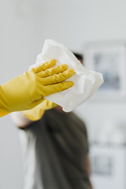 A person wearing a bright blue cleaning glove and a yellow sleeve holding a black dustpan brush with stiff bristles, poised to sweep a smooth white surface. The background features a plain light-color